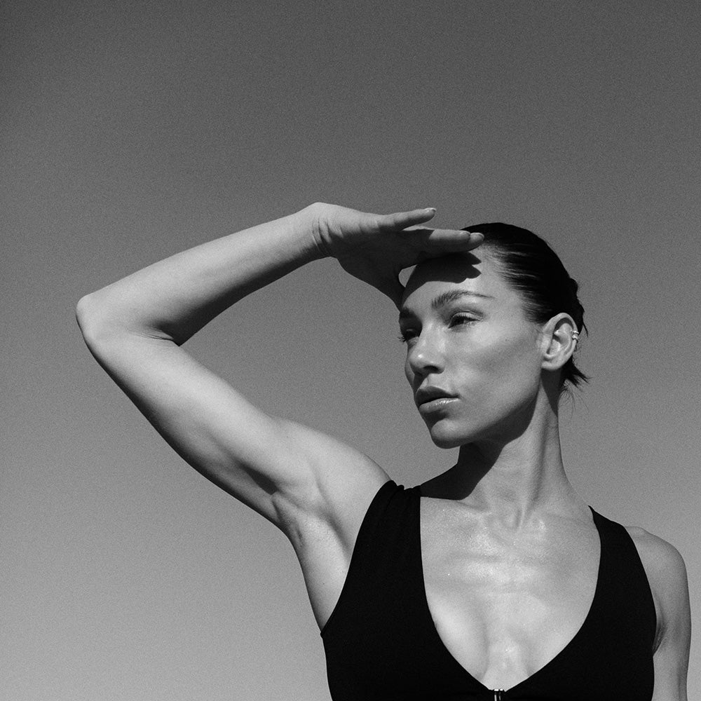 Black and white portrait of a woman in a sleeveless top shielding her eyes from the sun against a clear sky.